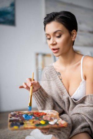 Sensual artist holding paintbrush and palette in studio 