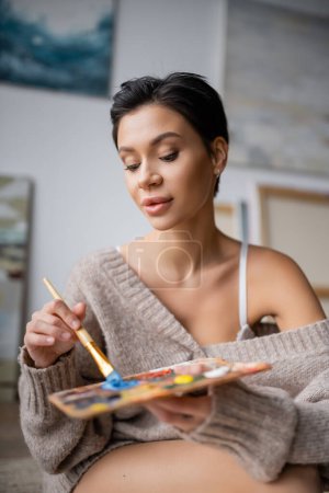 Seductive brunette artist in sweater mixing paints on palette in workshop 