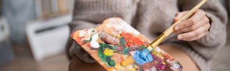 Téléchargez les photos : Cropped view of artist in sweater holding paintbrush and palette in workshop, banner - en image libre de droit