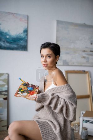 Sensual artist in sweater holding palette and paintbrush in workshop 