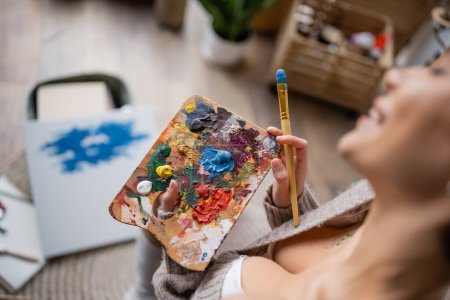 Foto de High angle view of sensual artist holding palette with pints and paintbrush in workshop - Imagen libre de derechos