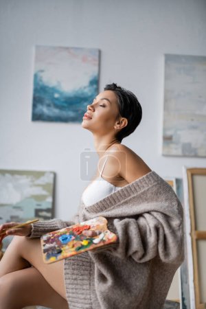 Sensual artist holding blurred palette and paintbrush in workshop 