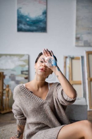 Tattooed artist in sweater holding hand in paint near face in workshop 