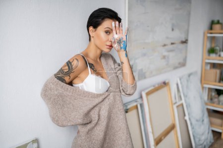 Tattooed artist in bra and sweater holding hand in paint near face in studio 