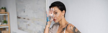 Photo for Seductive artist holding hand in paint near face and looking at camera in workshop, banner - Royalty Free Image
