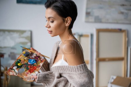 Sexy artist in bra and sweater holding palette and paintbrush