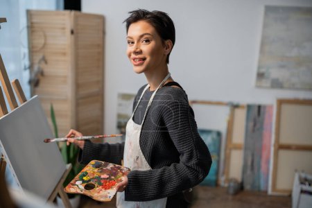 Smiling artist looking at camera while painting on canvas in studio 