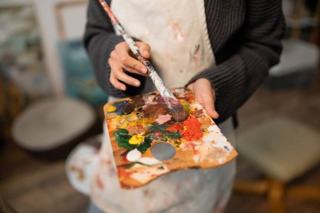 Téléchargez les photos : Cropped view of blurred artist in apron holding palette and paintbrush - en image libre de droit
