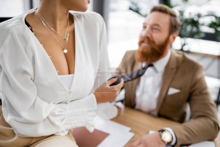 Photo for Cropped view of african american woman with big breast sitting on desk and pulling tie of bearded coworker in office - Royalty Free Image