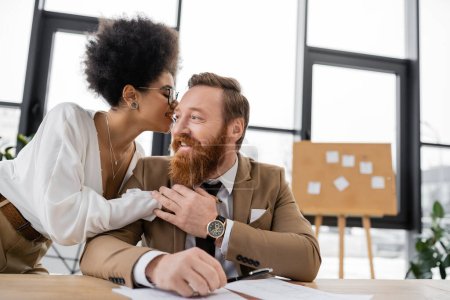 smiling african american businesswoman hugging while whispering in ear of cheerful coworker in office 