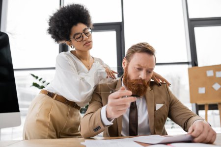 curly african american businesswoman touching shoulders of businessman working with documents in office 