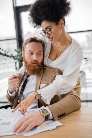 passionate african american woman leaning on shoulders of bearded man while pointing at charts 