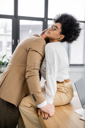 bearded man kissing neck of african american woman in glasses sitting on desk in office 
