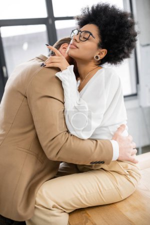 passionate man kissing neck of african american woman in glasses sitting on desk in office 