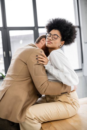 passionate man kissing neck of curly african american woman in glasses sitting on desk in office 