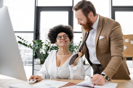 smiling african american woman pulling tie of bearded businessman in office 