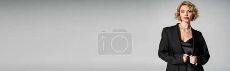 Photo for Curly young woman in black slip dress and blazer looking at camera isolated on grey, banner - Royalty Free Image