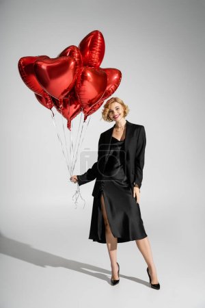 full length of happy blonde woman in black slip dress and blazer standing with red heart-shaped balloons on grey 