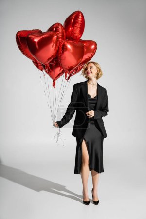 full length of cheerful woman in black slip dress and blazer looking at red heart-shaped balloons on grey 
