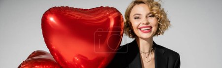happy young woman in black blazer smiling near red heart-shaped balloons isolated on grey, banner 
