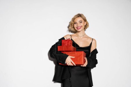 happy woman in black blazer and satin slip dress holding wrapped red presents on grey background 