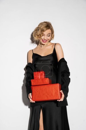 joyful woman in black blazer and satin slip dress holding wrapped red presents on grey 