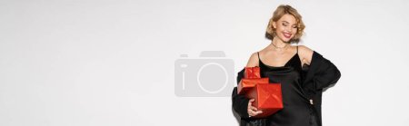 happy woman in black blazer and satin slip dress holding red presents while posing with hand on hip on grey, banner 