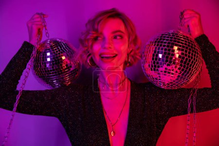 happy young woman with blonde hair standing in black dress and holding chains with disco balls on purple and pink 