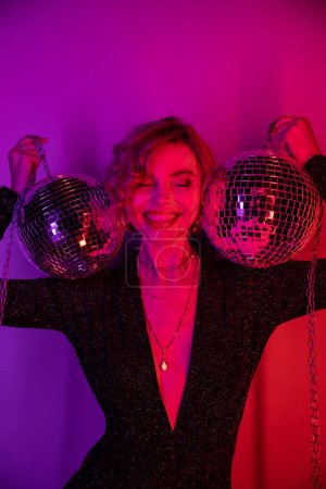cheerful young woman with blonde hair standing in black dress and holding chains with disco balls on purple and pink 