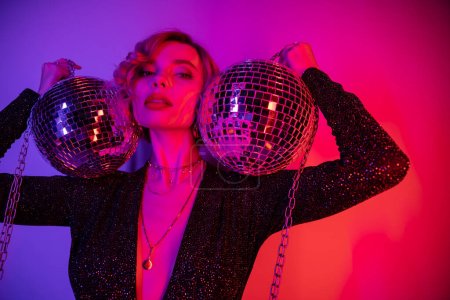Photo for Young woman with blonde hair standing in black dress and holding chains with disco balls on purple and pink - Royalty Free Image