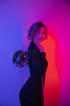 Foto de Young woman with blonde hair standing in tight dress and holding chain with disco ball on purple and pink - Imagen libre de derechos
