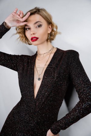 young woman with blonde curly hair and red lips posing in black shiny dress on grey 