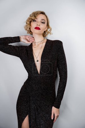 young woman with blonde hair and red lips posing in black shiny dress on grey 