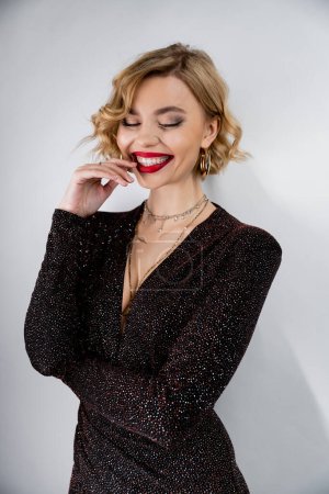 cheerful young woman with blonde curly hair and red lips posing in black shiny dress on grey 