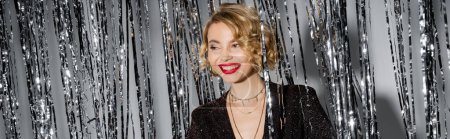 Foto de Positive young woman with red lips standing in black dress near shiny tinsel curtain on grey, banner - Imagen libre de derechos