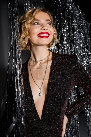 Photo for Joyful young woman in black dress smiling near shiny tinsel curtain on grey - Royalty Free Image