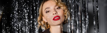 Foto de Blonde young woman with red lips looking at camera near shiny tinsel curtain on grey, banner - Imagen libre de derechos