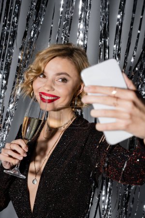 happy woman taking selfie while holding glass of champagne near tinsel curtain on grey
