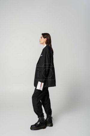 Photo for Side view of trendy woman posing in black oversize pantsuit and rough boots on grey background - Royalty Free Image
