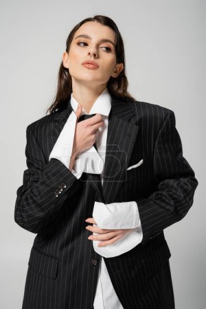 trendy woman in oversize shirt and blazer adjusting black tie while looking away isolated on grey