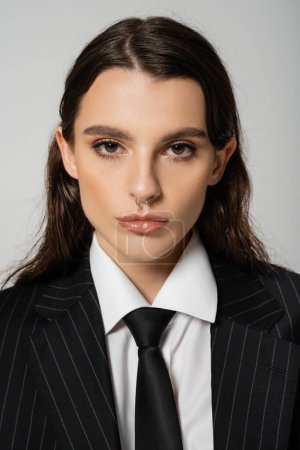portrait of brunette woman with makeup and piercing posing in stylish clothes and tie isolated on grey
