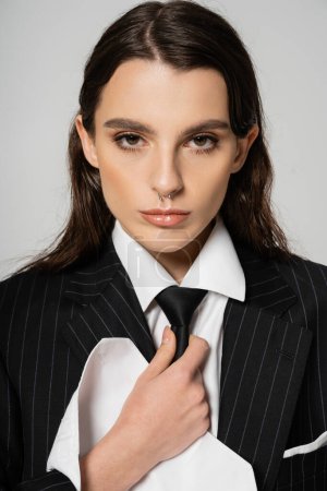 portrait of stylish brunette woman in black blazer and white shirt touching tie and looking at camera isolated on grey