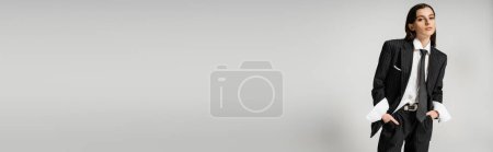 Téléchargez les photos : Brunette woman in fashionable oversize suit holding hands in pockets and looking at camera isolated on grey, banner - en image libre de droit