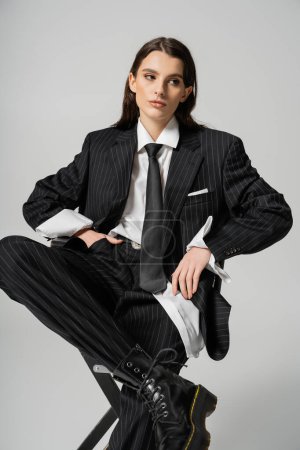stylish brunette woman in elegant oversize suit sitting with hand in pocket while looking away isolated on grey