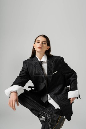 trendy woman in oversize formal wear sitting and looking at camera isolated on grey