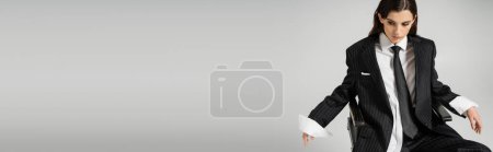 Téléchargez les photos : Young woman in white shirt and black blazer with tie looking down while sitting isolated on grey, banner - en image libre de droit