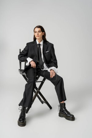 Photo for Full length of trendy woman in oversize formal wear and laced-up boots looking at camera while sitting on grey - Royalty Free Image
