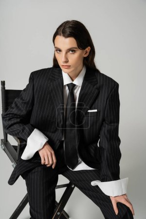 Photo for Stylish brunette woman in black striped suit and tie posing on chair and looking at camera isolated on grey - Royalty Free Image
