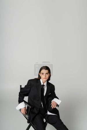 Téléchargez les photos : Trendy woman in oversize formal wear sitting and looking at camera isolated on grey with copy space - en image libre de droit
