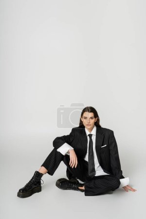full length of fashionable woman in black elegant suit and rough boots sitting on grey background